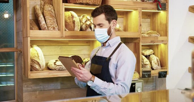 Young handsome man vendor in apron standing at the counter in bakery shop indoors tapping and typing on tablet at work. Portrait of Caucasian male baker in medical mask in bakehouse. Small business