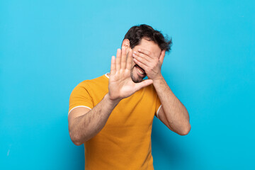 young handsome man covering face with hand and putting other hand up front to stop camera, refusing...