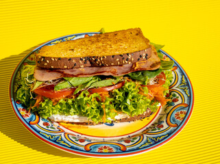 Exquisite ham sandwich with whole wheat bread, tomato, lettuce, avocado and cheese inside on a plate on a yellow table.