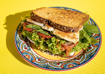 Delicious chicken sandwich with whole wheat bread, tomato, ham, lettuce and cheese inside on a plate on a yellow table.