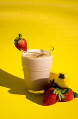 Delicious strawberry and banana shake inside a glass with strawberries and a banana on the side reflecting a black shadow on a yellow background.