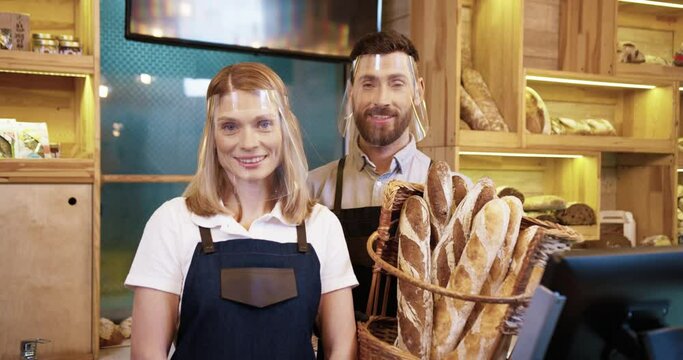 Portrait of Caucasian happy young married couple bakers in protective face shields and gloves holding basket with fresh bread standing in small own bakehouse in quarantine. Family business concept