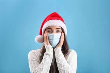 Beautiful emotional woman wearing Santa Claus hat and medical mask on light blue background