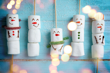 Cute snowmen made of marshmallows on light blue wooden background, flat lay. Bokeh effect