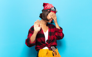young latin woman covering face with hand and putting other hand up front to stop camera, refusing...
