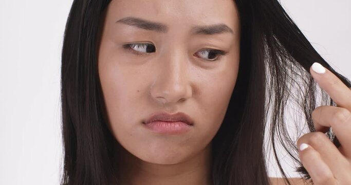 Close up portrait of young asian woman unhappy with her hair condition, looking sadly at camera, white studio background