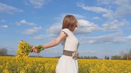 Fototapeta premium Happy girl in a white dress with a bouquet of rapeseed flowers in a yellow rapeseed field.