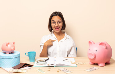 young latin woman looking happy, proud and surprised, cheerfully pointing to self, feeling...