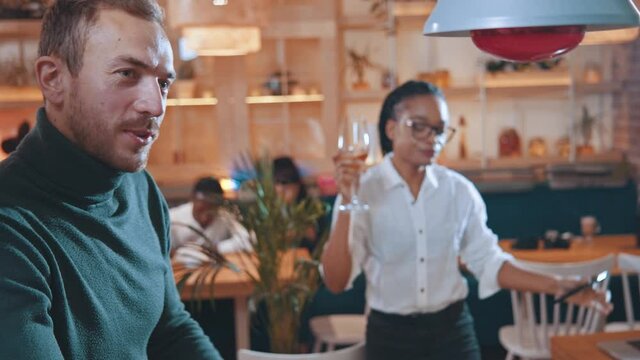 Young man holding her drunk afro-american funny girlfriend drinking lots of wine while staying in the cafe. Alcoholic woman. Humor concept.