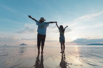 Fototapeta premium Silhouette of asian mother and daughter holding hand together standing on the beach looking at the beautiful sea and sky at the sunset time.