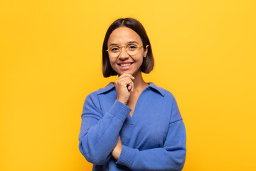 young latin woman looking happy and smiling with hand on chin, wondering or asking a question,...