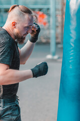 Street Fighter in Black Clothes and Bandages on the Wrist Boxing in Punching Bag Outdoors. Young Man Doing Box Training and Practicing His Punches at the Outside Gym