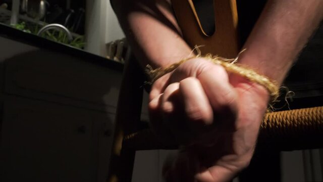 Man struggles with hands tied to chair with rope in kitchen at night