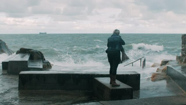 A woman standing by the ocean and taking pictures of the waves breaking