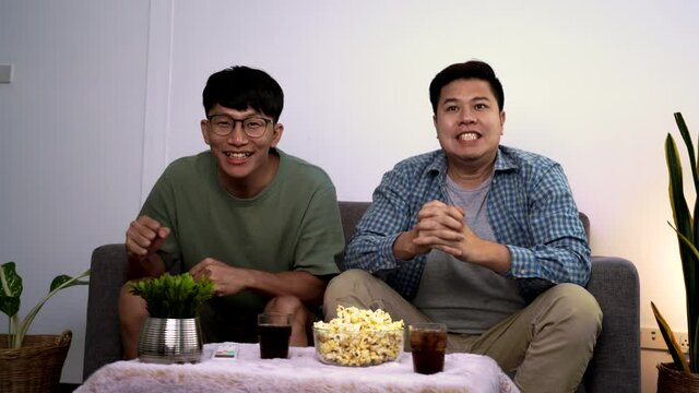 Asian friends or football fans sitting on sofa watching soccer or football game on tv at home, sports and entertainment concept.
