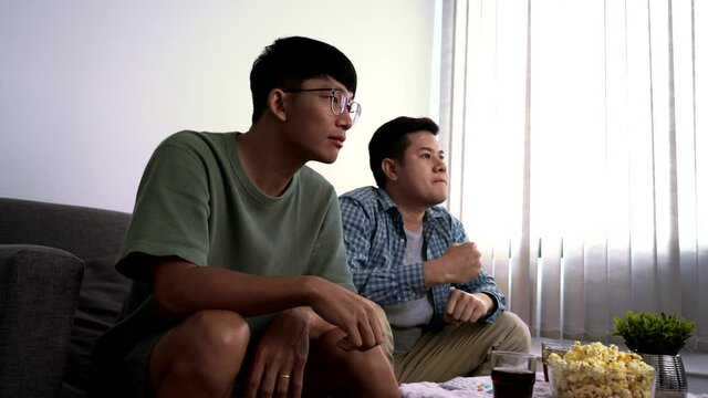 Asian friends or football fans sitting on sofa watching soccer or football game on tv at home, sports and entertainment concept.
