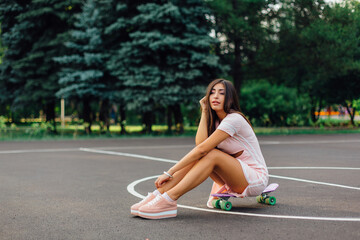 Portrait of a smiling charming brunette female sitting on her skateboard on a basketball court.