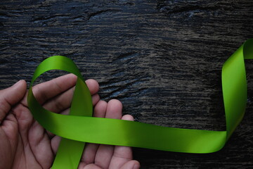 Top view of hands holding lime green color ribbon on dark background. Non-hodgkin lymphoma cancer, lyme disease, muscular dystrophy and postpartum depression awareness concept