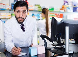 Smiling man pharmacist is writing to note inventsry of medicines near computer in drugstore