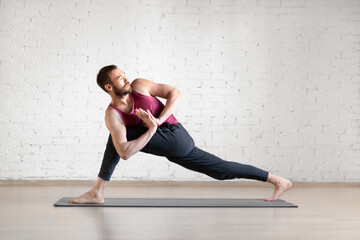 Male yoga, practice indoor. Caucasian lean man doing deep lunge with spine twist exercise on grey mat in fitness loft studio, selective focus.