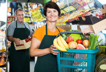 adult smiling female seller is offering basket with goods in the market.
