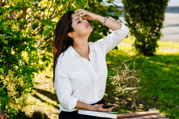 Executive woman sitting with a book using the phone on a park bench.