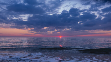 Evening seascape. Shades of blue and pink. There are picturesque clouds in the sky. The sun is low above the water. There are small waves and foam on the shiny surface of the sea.