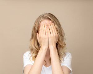 Faceless closeup portrait woman covering hand on face, filling with deep sadness. Isolated on beige wall. Pretty female with curly hair in white t shirt