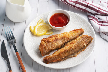 Fried fish hake Pollock with tomato sauce and slices of fresh lemon. Concept eating fatty fast food. White wooden table with Copy space.