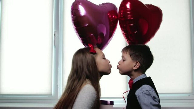 Boy hands a box with a gift to a girl on the background of balloons in the shape of hearts. Two happy Caucasian children kiss with their lips stretched out. The concept of Valentine's day