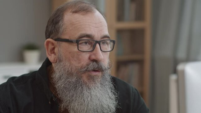 Close-up Of Elderly Caucasian Man With Heavy Beard And Glasses Talking To Invisible Partner In Office