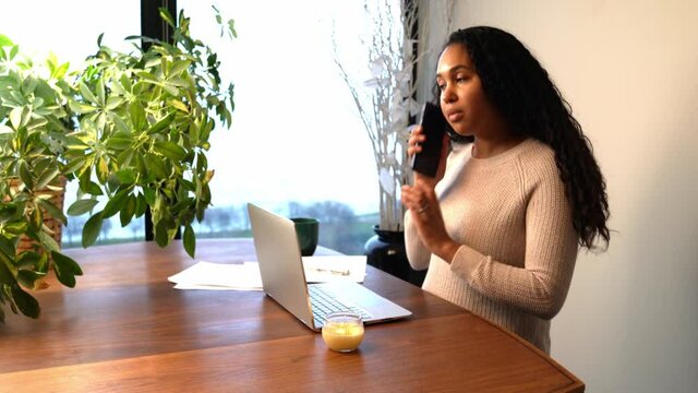 Beautiful African American Woman Types On Her Laptop Computer Before Picking Up Her Phone To Take A Call At Her Remote Work From Home Station At Her Dining Room Table With Coffee Cup And Papers.