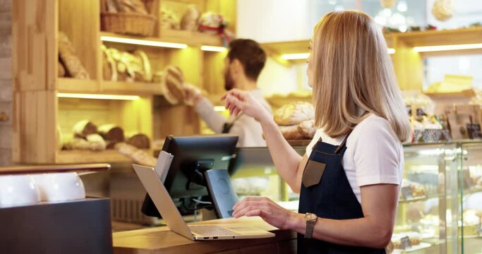 Close up of beautiful young Caucasian woman seller in apron standing in bakery, tapping and typing on laptop talking to male colleague working on background. Bakehouse concept. Small business