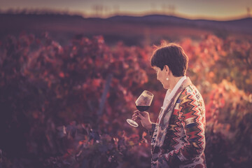 A woman with a glass of red wine stands near a vineyard in the autumn at sunset. Wine tour