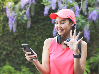 healthy Asian woman in pink sportswear standing in the garden with purple flowers, wearing headphones, using mobile phone to make video call and waving to the screen. sports and technology   concept.