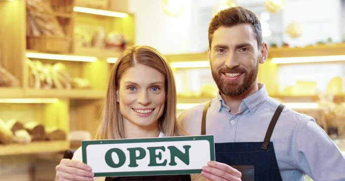 Close up of cheerful Caucasian young male and female bakers standing in own bakery and holding Open sign in hands, looking at camera and smiling. Bakehouse reopening. Small business opening