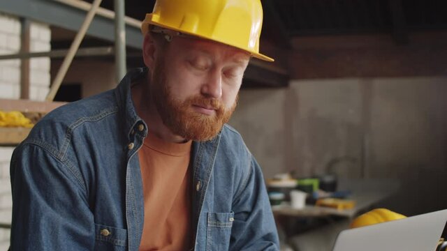 Tilt up shot of bearded male architect in hardhat using laptop and then posing for camera while working at construction site