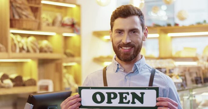 Close up of happy Caucasian bearded handsome male baker standing in own bakery and holding Open sign in hands, looking at camera and smiling. Bakehouse reopening. business opening. Bakehouse concept