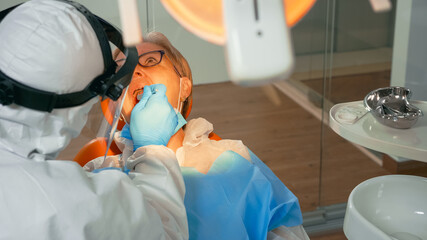 Woman on dental chair in dentist surgery treated with dental drill during covid-19 pandemic. Orthodontist lighting the lamp wearing face shield, protection suit, mask and gloves talking with patient