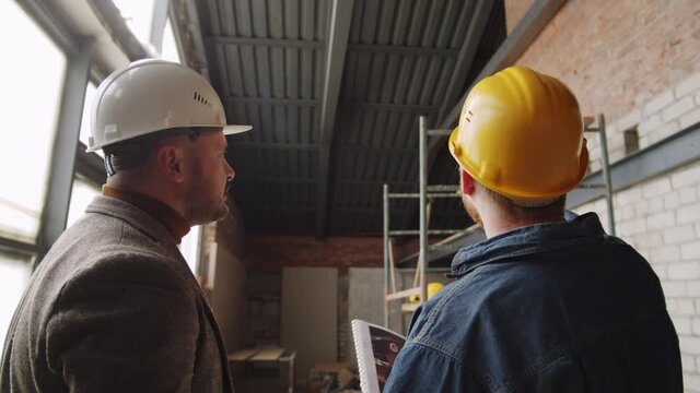Back view of two male engineers in hardhats discussing ceiling construction while working together on building new residential house