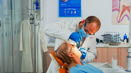 Young man orthodontist examining senior patient using sterile dental tools in stomatological clinic. Nurse lighting the lamp, doctor speaking to patient sitting on stomatological chair.