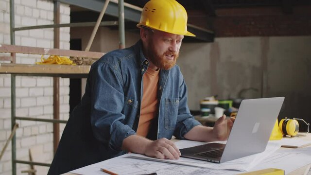 Caucasian male architect in hardhat speaking via online video call on laptop while working at construction site