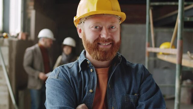 Chest up portrait shot of happy bearded male architect in hardhat standing at construction site, looking at camera and smiling
