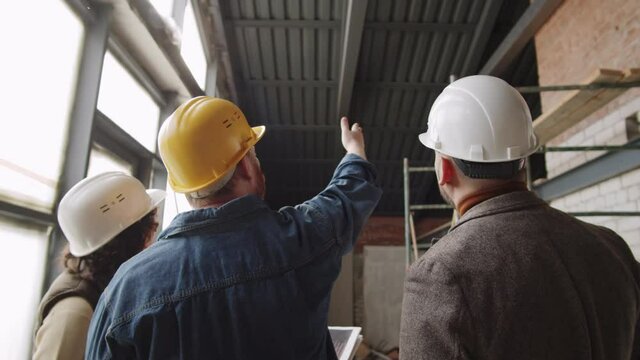 Back view of team of male and female architects in hardhats pointing at ceiling construction in new unfinished apartment and having discussion during workday