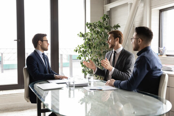 Young successful male business partners or colleagues sit gather at meeting in office, discuss cooperation partnership together. Confident multiracial businessmen talk brainstorm at briefing.