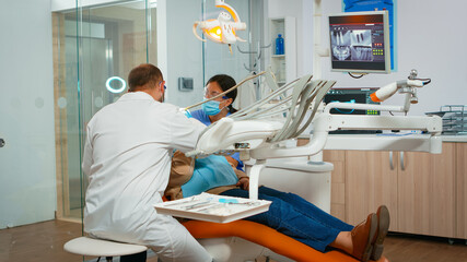 Dentist treating teeth to senior woman patient in clinic lying on chair with open mouth. Doctor and nurse working together in modern stomatological office wearing protection mask