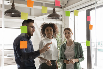 Young diverse businesspeople work attack sticky notes on glass wall in boardroom develop business...
