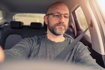 Bald attractive male driver in a car. The model in his 40s, wearing grey shirt. Grey and dark beard, metal glasses. Selective focus. Warm tones