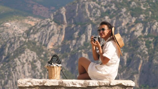 Young woman sitting on stone bench at Delphi landscape with mountains from high viewpoint at sunset, Taking pictures. Greece. Fashion white dress, sunglasses, large hat.