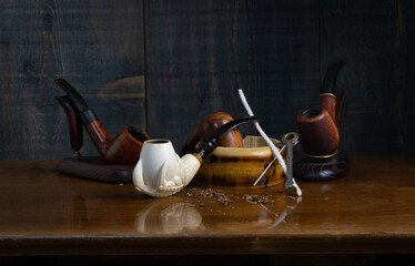 Composition with smoking pipes: in the foreground a pipe made of sea foam (meerschaum), two of heather and one of cherry, an ashtray, pipe cleaning accessories and tobacco crumbs.